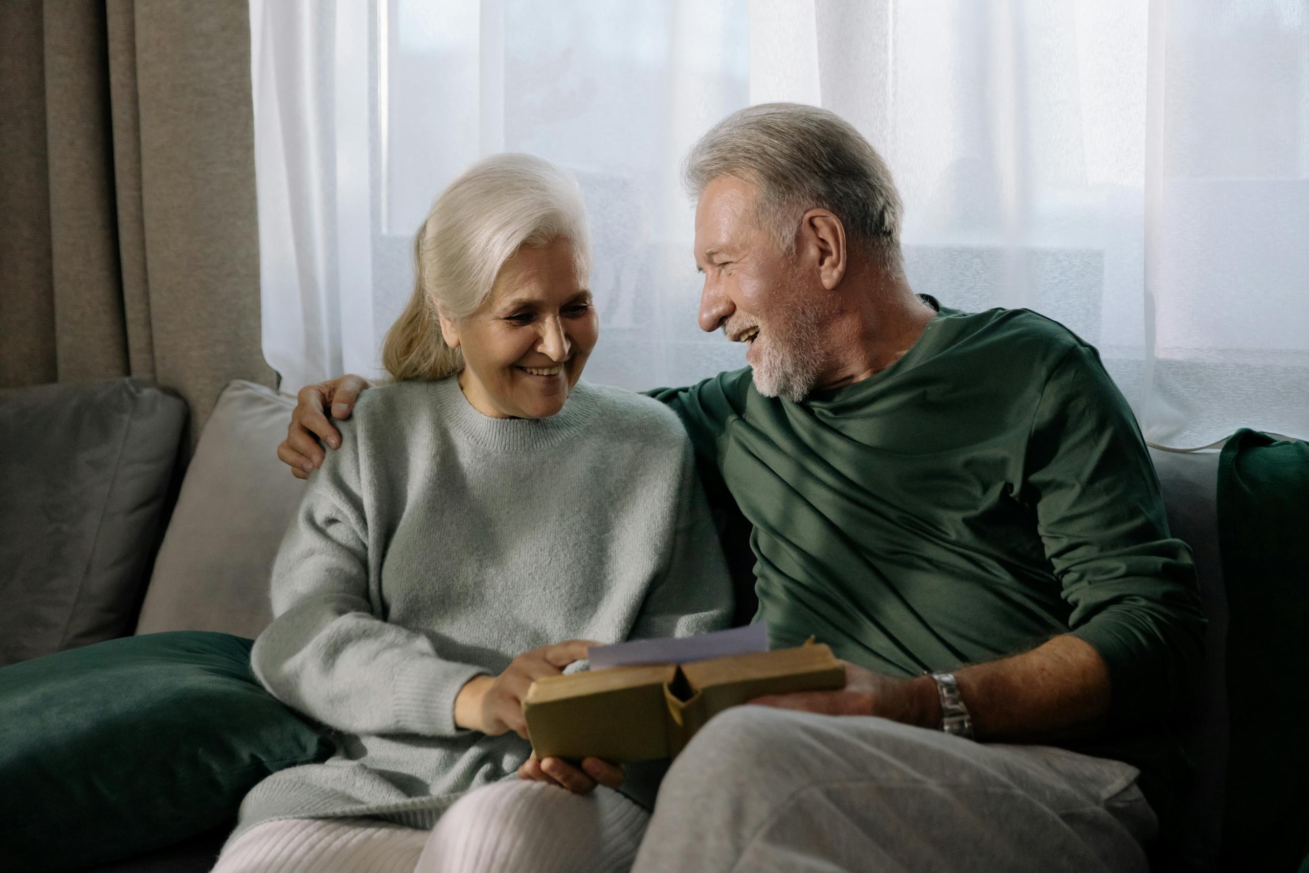 Senior couple embracing and reading a book on a sofa, sharing a joyful and intimate moment indoors.